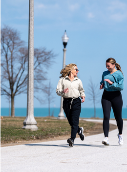Two women jogging together, smiling, on a paved path near a lake.